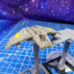 May include: A detailed model of a spacecraft, painted in shades of gray with yellow accents on the wings. The model is mounted on a gray stand, showcasing its intricate design. A second, unpainted model is also visible.
