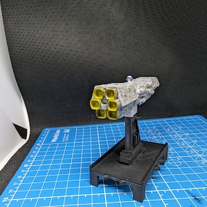 May include: A white and gray model spaceship with yellow engine details. The spaceship is mounted on a black stand with a square base.