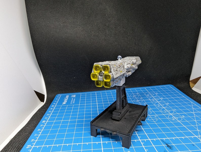 May include: A white and gray model spaceship with yellow engine details. The spaceship is mounted on a black stand with a square base.