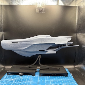 May include: A gray, unpainted 3D-printed model of a futuristic spaceship featuring a sleek, elongated hull with intricate paneling details, mounted on a black display stand. The vessel has a pointed bow and a complex, layered rear section.