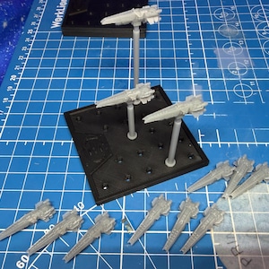 Puede incluir: Una colección de modelos de naves espaciales grises en miniatura, algunos montados sobre soportes, dispuestos sobre una esterilla de corte azul con cuadrícula. Los modelos presentan detalles de alas y casco.