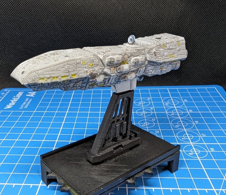 May include: A white and gray model spaceship with yellow details, mounted on a black display stand. The spaceship is on a blue grid surface.