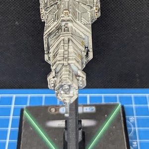 May include: A gray and white spaceship model with a black base. The spaceship has a long, cylindrical body with a pointed nose and a series of windows and hatches along its sides. The base has a green X pattern on it.