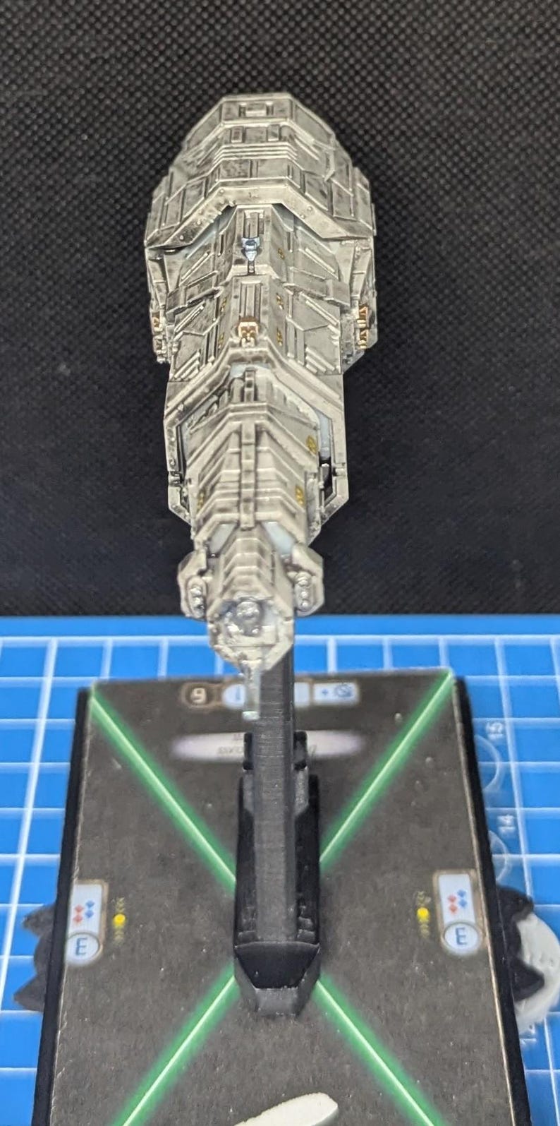 May include: A gray and white spaceship model with a black base. The spaceship has a long, cylindrical body with a pointed nose and a series of windows and hatches along its sides. The base has a green X pattern on it.
