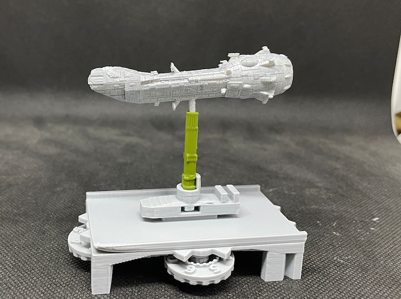 May include: A gray 3D printed model of a spaceship on a gray 3D printed base with a green support rod. The spaceship is long and narrow with a rounded front and a pointed back. The base has a circular platform with a gear on the bottom. The support rod is attached to the base and the spaceship.
