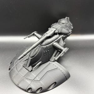 May include: A grey 3D-printed model of a futuristic vehicle. The model has a rounded base with multiple circular openings, a central cockpit area, and extended arm-like structures with detailed components. The design suggests a spacecraft or sci-fi vehicle.