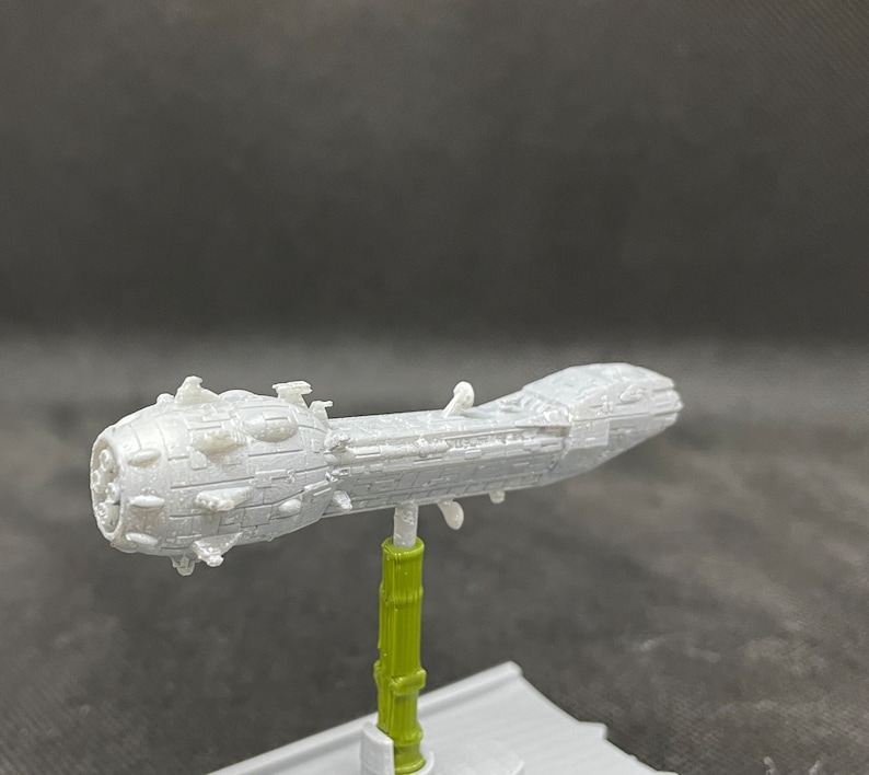 May include: A white 3D printed model of a spaceship with a long, cylindrical body and a rounded, spiked front. The model is mounted on a green stand.