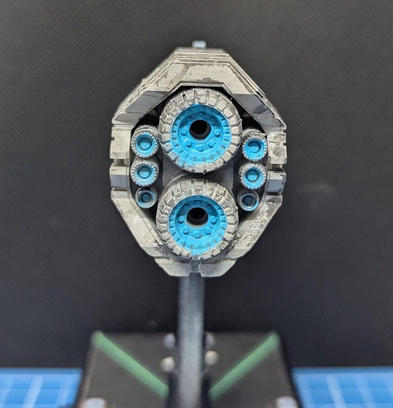 May include: A close-up of a model spaceship with a gray and silver paint finish. The spaceship has multiple blue exhaust ports on the back.