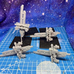 Puede incluir: Cuatro modelos grises en miniatura de naves espaciales se exhiben sobre una superficie de cuadrícula azul, cada uno montado sobre una base negra. El fondo presenta una tela estrellada azul y púrpura. Los modelos tienen diseños geométricos intrincados.