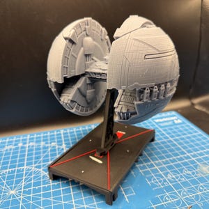 May include: A grey 3D printed model of a spherical space station featuring intricate panelling and mechanical details. The model is mounted on a black display stand with red accent lines, positioned on a blue cutting mat with grid markings.