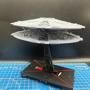 May include: A grey 3D-printed model of a circular spaceship with two stacked, detailed disc-shaped sections. The model is mounted on a black vertical stand attached to a rectangular black base featuring red lines and the text "MC80" in white.