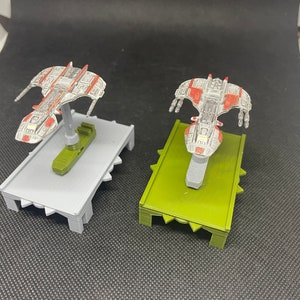 May include: Two miniature spaceships painted in white, red, and gray, with one on a gray base and the other on a green base.