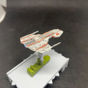 May include: A white and red spaceship model with a gray base. The spaceship has a long, narrow body with a pointed nose and two wings. The wings have a red stripe running along the top edge. The base is a simple, flat platform.