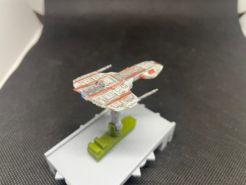 May include: A white and red spaceship model with a gray base. The spaceship has a long, narrow body with a pointed nose and two wings. The wings have a red stripe running along the top edge. The base is a simple, flat platform.