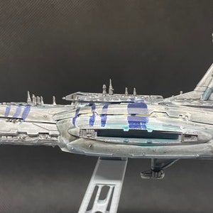 Puede incluir: Un modelo detallado de una gran nave espacial gris con marcas azules y amarillas. La nave espacial tiene un cuerpo largo y estrecho con un gran motor rectangular en la parte trasera. El modelo est&aacute; hecho de pl&aacute;stico y est&aacute; pintado en un estilo realista.