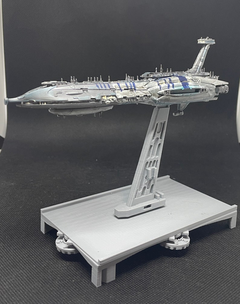 Puede incluir: Un modelo gris y azul de una nave espacial en un soporte gris. La nave espacial tiene un cuerpo largo y delgado con una nariz puntiaguda y un ala grande y plana en la parte superior. El soporte es de pl&aacute;stico y tiene una base rectangular.