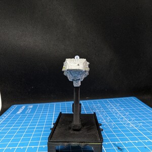 May include: A white and gray miniature spaceship model on a black stand. The model has a detailed design with a rounded top and a pointed bottom.