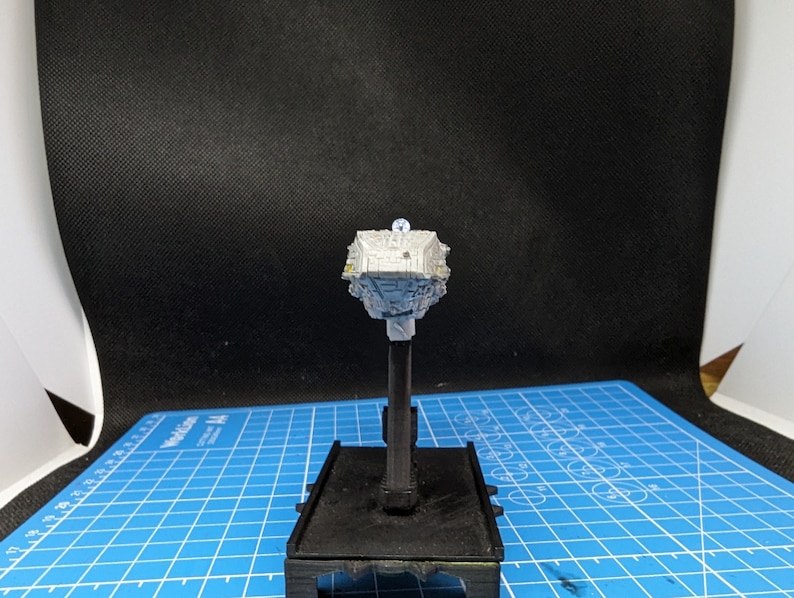 May include: A white and gray miniature spaceship model on a black stand. The model has a detailed design with a rounded top and a pointed bottom.