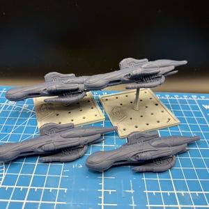 May include: Four gray spaceship models are displayed on a blue cutting mat. Two models are elevated on gray stands, while two others rest directly on the mat. The spaceships have a sleek, futuristic design.