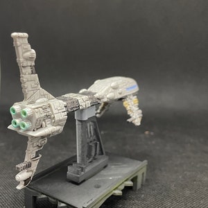 May include: A gray and black spaceship model with a stand. The spaceship has a long, thin body with a large engine at the back. The engine has four green exhaust ports. The spaceship is on a black stand with a gray base.