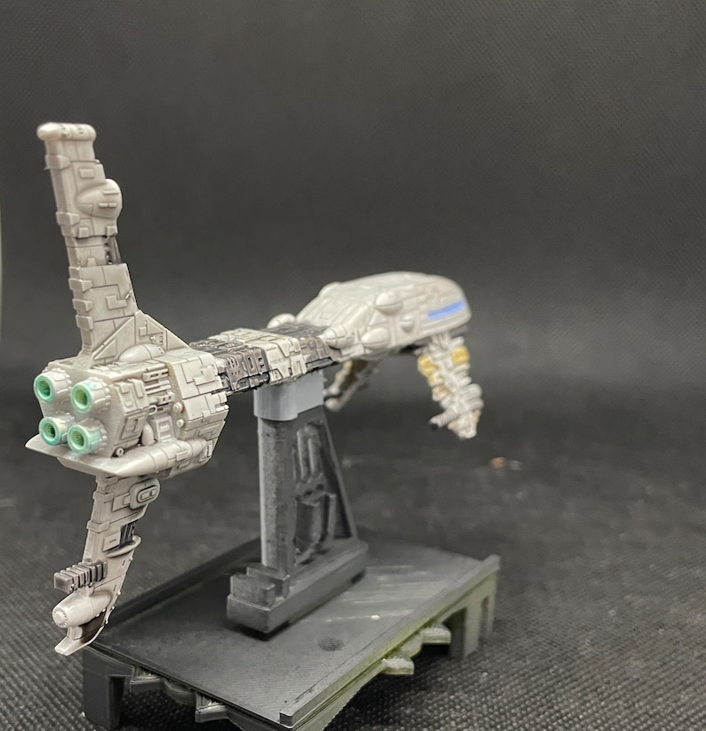 May include: A gray and black spaceship model with a stand. The spaceship has a long, thin body with a large engine at the back. The engine has four green exhaust ports. The spaceship is on a black stand with a gray base.