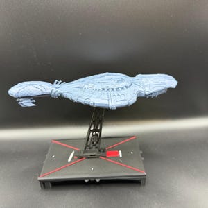 Puede incluir: Maqueta de nave espacial azul claro con detalles intrincados, montada sobre un soporte negro con detalles rojos. El modelo tiene un diseño aerodinámico, sugiriendo un tema futurista. La base es cuadrada con líneas rojas.