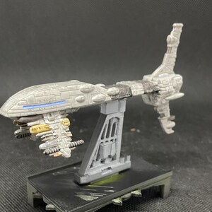 May include: A gray and white spaceship model with a long, thin body and a large, round cockpit. The spaceship has a variety of details, including a small, rectangular window on the side of the cockpit and a series of small, round windows on the underside of the body. The spaceship is mounted on a gray stand.