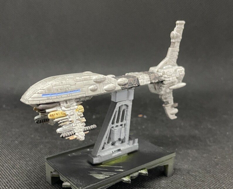 May include: A gray and white spaceship model with a long, thin body and a large, round cockpit. The spaceship has a variety of details, including a small, rectangular window on the side of the cockpit and a series of small, round windows on the underside of the body. The spaceship is mounted on a gray stand.