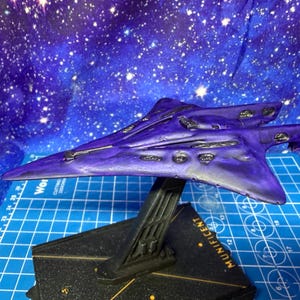 May include: A purple spaceship model with a sleek design, mounted on a black stand. The model has detailed features and is set against a starry blue backdrop. The base of the stand has the word "MUNIFICENT" printed on it.
