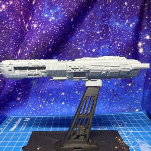 Puede incluir: Maqueta gris de nave espacial, de unos 30 cm de largo, expuesta sobre un soporte negro sobre un fondo estrellado. El modelo tiene detalles de superficie y es probablemente un artículo de colección. El soporte está sobre una esterilla de corte azul.