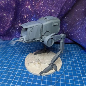 Puede incluir: Modelo AT-ST gris impreso en 3D, un caminante bípedo de una franquicia de ciencia ficción. El modelo está sobre una base circular con una superficie texturizada. El fondo es una tela azul y morada.