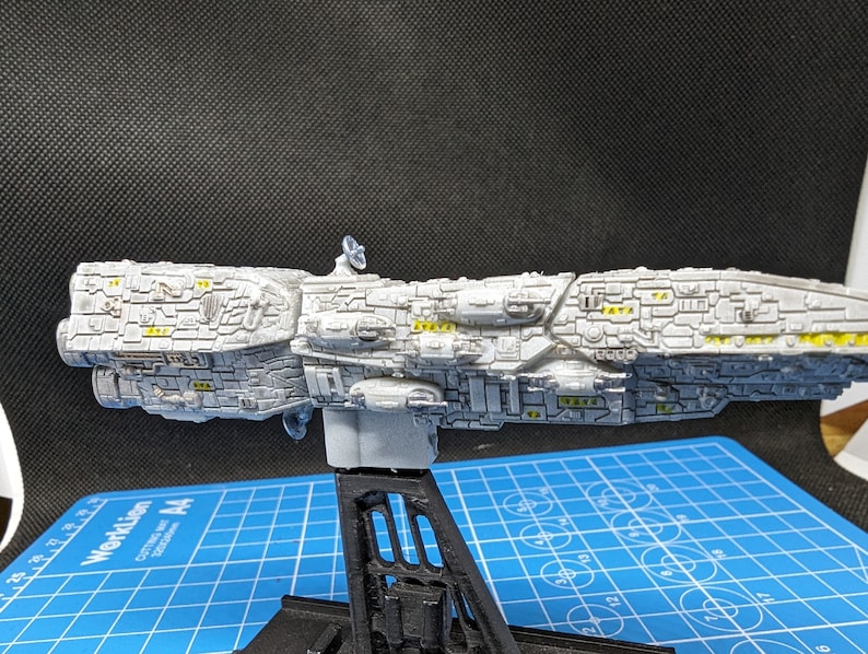 May include: A white model spaceship with yellow details, mounted on a black stand. The spaceship is detailed with a variety of textures and shapes.