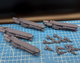 May include: A collection of gray miniature spaceship models, including several larger capital ships and smaller fighter craft. The models are arranged on a blue cutting mat with grid lines, suggesting a hobby or gaming context.