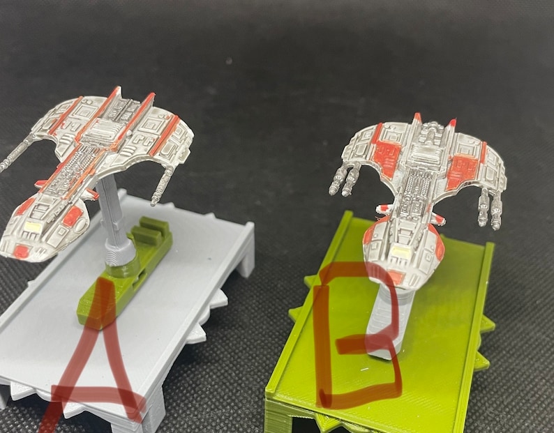 May include: Two miniature spaceships, one on a gray base and one on a green base, both with red and white details. The spaceships are on display stands.