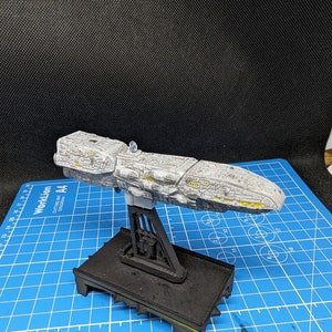 May include: A gray and white spaceship model with yellow accents, mounted on a black stand. The model is on a blue cutting mat with white markings.