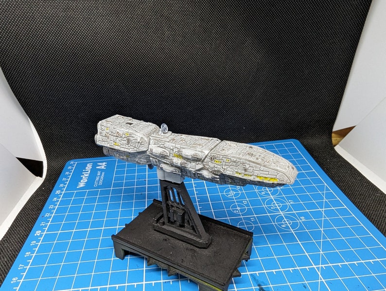 May include: A gray and white spaceship model with yellow accents, mounted on a black stand. The model is on a blue cutting mat with white markings.