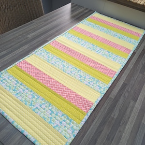 May include: A striped table runner with alternating bands of yellow, pink, and light blue fabric. The fabric has a subtle floral pattern.