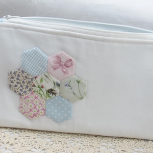 May include: White zippered pouch with a patchwork design of hexagonal fabric pieces in shades of blue, pink, and white. The fabric pieces feature floral patterns, gingham, and a pink bow.