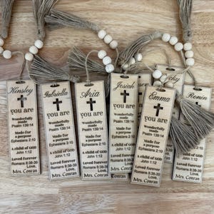 Op de afbeelding: Houten bladwijzers met kwastjes en namen als Kinsley, Gabriella en Aria. Elke bladwijzer heeft een kruis en inspirerende tekst uit de Bijbel. De bladwijzers zijn verbonden met een kralenketting.