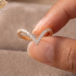 May include: A gold-colored V-shaped ring with small, clear gemstones set along the band. The ring is held between fingers, showcasing its delicate design. The ring is part of a jewelry collection.