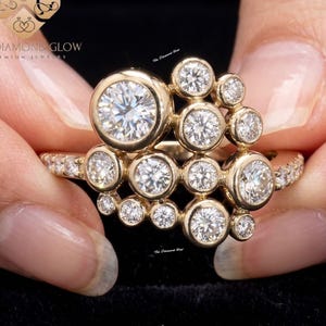 May include: A gold ring featuring a cluster of round-cut diamonds. The ring has a unique design with a large central diamond surrounded by smaller stones. The band is also adorned with smaller diamonds. The ring is held against a black background.