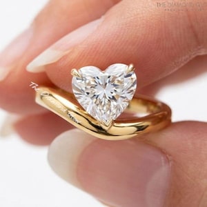May include: A gold ring featuring a large, heart-shaped diamond. The ring's band curves gracefully, cradling the sparkling gemstone. The diamond's facets catch the light, creating a brilliant display. The ring is held by a person.