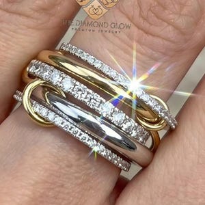 Può includere: Un anello a più fasce che presenta una combinazione di bande in oro giallo lucido e metallo color argento. Diverse bande sono incastonate con piccole pietre chiare e scintillanti. Il design è composto da anelli metallici intrecciati.