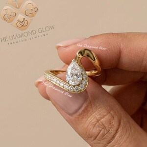 May include: A gold ring featuring a pear-shaped center stone and a curved band accented with small round stones. The text THE DIAMOND GLOW PREMIUM JEWELRY is visible in the upper left corner, along with a logo featuring four circular icons.