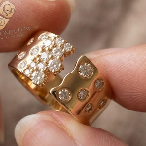 May include: A gold open-style ring featuring a wide band with a polished finish. One side of the ring is densely set with multiple small round clear stones, while the other side features a scattered arrangement of larger round clear stones.