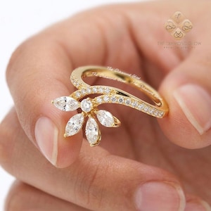 May include: A gold ring featuring a floral design with marquise-cut diamonds and smaller round diamonds. The ring has a curved band and a cluster of diamonds forming a flower shape. The ring is held in a hand.