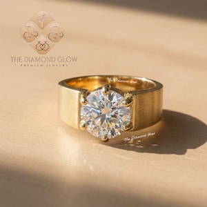 May include: A gold ring with a large, round, clear diamond center stone. The ring has a wide, brushed gold band. The Diamond Glow premium jewelry logo is visible in the background.