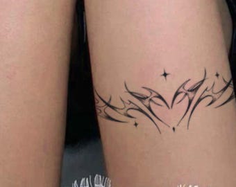 May include: Black tattoo design on the thigh featuring a heart shape with star accents. The tattoo is a continuous line design with sharp, angular elements. The skin tone is light, and the tattoo is placed above a white lace trim.