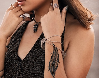 May include: A black feather temporary tattoo on a person's wrist. The tattoo is a simple design with a few small dots around it. The word "TATNOW" is visible at the bottom of the image.