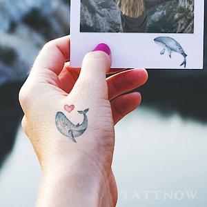 Può includere: Un tatuaggio temporaneo di una balena con un cuore rosso sulla schiena. Il tatuaggio è sulla mano di una persona.